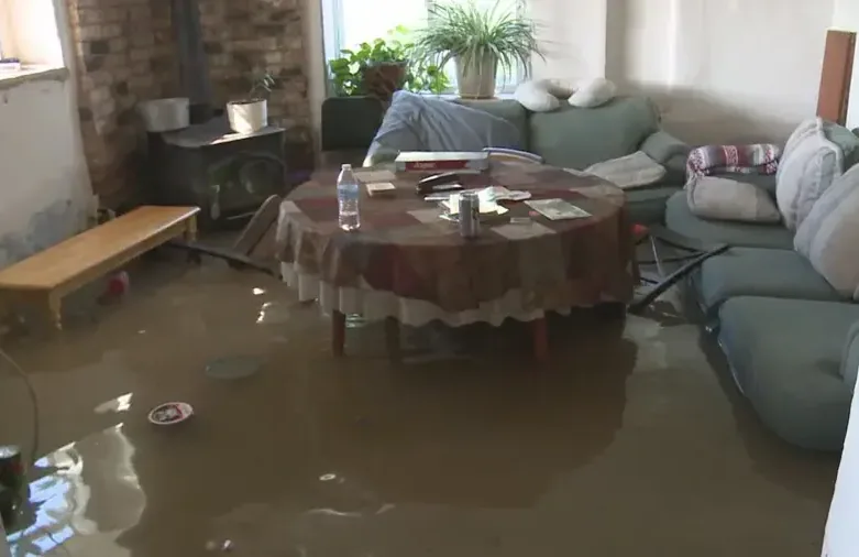 Muddy floodwater covers a furnished living room floor around chairs and a table
