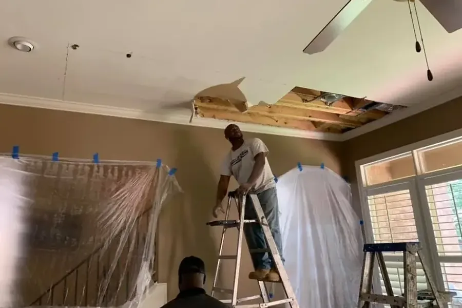 A worker on a ladder opens a damaged ceiling beneath exposed framing